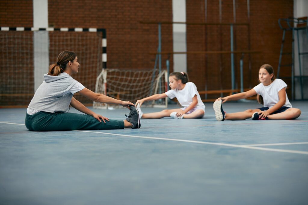 Female coach and elementary students stretching during physical education class at school gym.